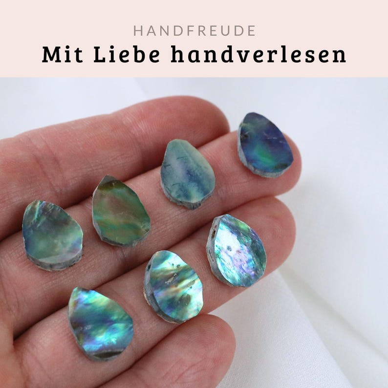 May include: Seven iridescent abalone shell pieces in various shades of green, blue, and purple. The pieces are shaped like teardrops and are arranged in a hand.