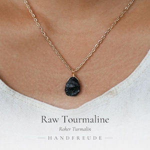 以下が含まれることがあります： ゴールドトーンのチェーンネックレスで、金色のワイヤーで固定された不規則な形の原石のブラックトルマリンペンダントが付いています。画像の下部には「Raw Tourmaline」、「Roher Turmalin」、「HANDFREUDE」という文字が黒色で印刷されています。