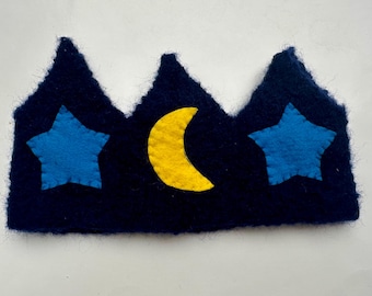 May include: A navy blue felt crown with three peaks. Two blue stars and a yellow crescent moon are sewn onto the crown. The crown is a whimsical accessory.