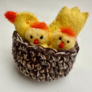 May include: A handmade felted Easter decoration featuring a yellow hen with two chicks nestled in a brown and beige crocheted nest. The chicks have orange crests and red beaks. The hen has an orange beak and a small yellow bead.