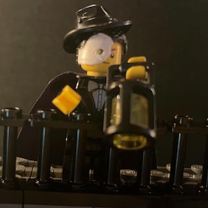 Op de afbeelding: Een Lego minifiguur draagt een zwart pak, een zwarte hoge hoed en een wit masker. De minifiguur houdt een zwarte lantaarn vast met een geel glazen venster. De minifiguur staat voor een zwart metalen hek.