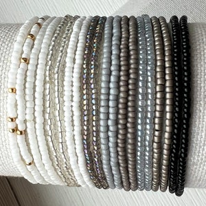 May include: A stack of 18 beaded bracelets in various shades of gray, white, and black. The bracelets are made with small, round beads and some have gold accents.