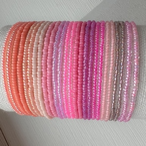 May include: A stack of colorful beaded bracelets in shades of pink, orange, and white. The bracelets are made with small, round beads and are arranged in a gradient pattern.