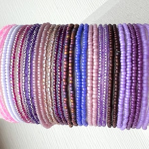 May include: A collection of twenty-two individual beaded bracelets arranged in a row. The bracelets feature small, round glass beads in a gradient of colors including light pink, lavender, magenta, deep purple, metallic bronze, and blue.