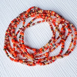 Peut inclure: Plusieurs brins de perles colorées en rouge, orange et or. Les perles sont petites et enfilées, créant un effet de superposition. Il s'agit probablement d'un bijou de taille ou d'un bracelet.