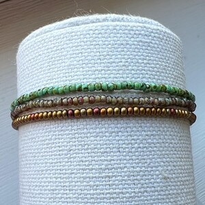 May include: A set of four beaded bracelets in shades of green, brown, and gold. The bracelets are stacked on a white fabric surface.