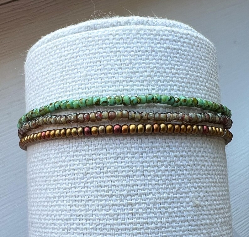 May include: A set of four beaded bracelets in shades of green, brown, and gold. The bracelets are stacked on a white fabric surface.