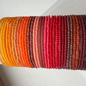 May include: A collection of beaded bracelets in a gradient of colors, from yellow-orange to deep burgundy. The bracelets are made of small, round beads and are displayed on a white cylindrical form.