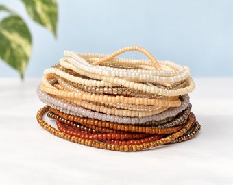 Puede incluir: Una pila de varias pulseras finas de cuentas con una paleta de colores en degradado. Las cuentas pasan del blanco crema y beige claro en la parte superior a tonos grises, marrones y rojo terracota oscuro en la base de la pila circular.