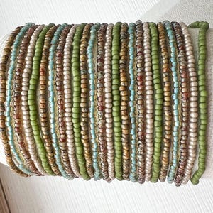 May include: A multi-strand bracelet composed of numerous rows of small seed beads in various shades of olive green, light blue, metallic bronze, and cream. The beads are arranged in a repeating pattern, creating a textured, cylindrical design.