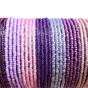 May include: A collection of multiple beaded bracelets arranged in a row, featuring a gradient color palette that transitions through various shades of purple, lavender, magenta, and soft pink, composed of small, uniform, round glass seed beads.