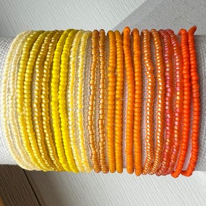 May include: A collection of twenty-one individual beaded bracelets arranged in a gradient color pattern. The beads transition from pale cream and bright yellow on the left to vibrant orange and deep reddish-orange on the right side.