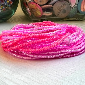 May include: A close-up of a stack of pink and purple seed beads. The beads are arranged in a graduated pattern, with the darkest pink beads at the bottom and the lightest purple beads at the top.