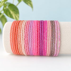 May include: A stack of multiple beaded bracelets featuring a gradient color scheme that transitions from bright orange and coral to various shades of pink, magenta, and purple, ending with translucent light purple and iridescent lavender beads.