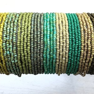 May include: A collection of beaded bracelets in various shades of green, yellow, and brown. The bracelets are made of small, uniform beads and are displayed stacked together. The color palette ranges from light yellow-green to dark olive green.