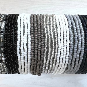 May include: A collection of 16 beaded bracelets in various shades of gray, white, and black. The bracelets are arranged in a row, with the darkest colors on the right and the lightest colors on the left.