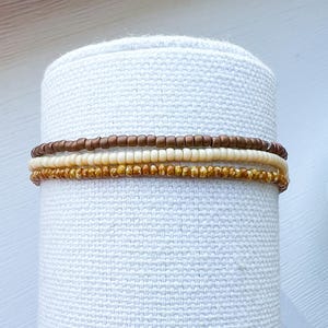 May include: Three beaded bracelets stacked on a white fabric surface. The top bracelet is brown, the middle bracelet is white, and the bottom bracelet is a speckled brown and gold.
