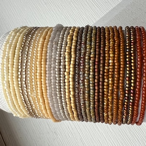 May include: A collection of twenty-one individual beaded bracelets arranged in a gradient color sequence. The bracelets feature small, round glass beads in shades ranging from cream and beige to tan, brown, bronze, and deep copper-toned orange.