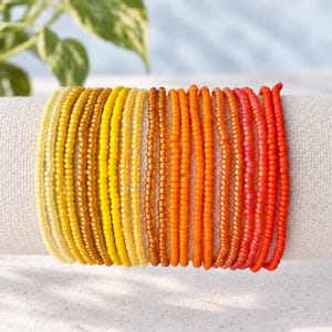 May include: A multi-strand bracelet featuring small, round glass beads arranged in a vibrant color gradient. The beads transition smoothly from pale yellow to golden yellow, deep orange, and finally to a bright, saturated red hue.