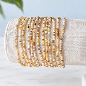 May include: A set of ten delicate beaded bracelets featuring a mix of small glass beads in shades of cream, translucent white, metallic gold, and soft amber. The bracelets are arranged side by side, showcasing a uniform, slender, and circular design.