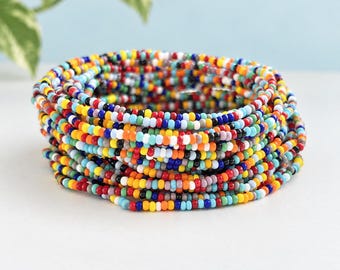 May include: A stack of multiple circular bracelets made from small, round, multi-colored glass seed beads. The beads feature a vibrant palette including red, orange, yellow, green, blue, white, and black, arranged in a repeating, random pattern.