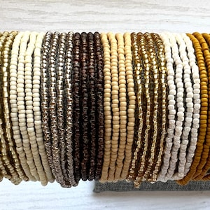 May include: A collection of beaded bracelets in various colors, including gold, cream, silver, brown, and mustard yellow. The bracelets are displayed stacked together, showcasing the different shades and textures of the beads. The bracelets are a fashion accessory.