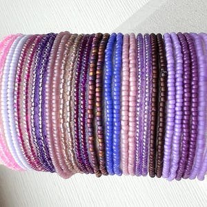 May include: A collection of twenty-two thin beaded bracelets arranged in a row. The bracelets feature small, round glass beads in various shades of pink, purple, lavender, violet, and deep plum, with some beads having a matte or metallic finish.