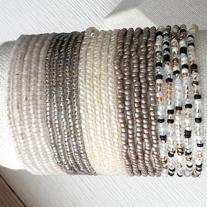 K&ouml;nnte beinhalten: Ein Stapel Perlenarmb&auml;nder in verschiedenen Wei&szlig;-, Grau- und Braunt&ouml;nen. Die Armb&auml;nder bestehen aus kleinen, runden Perlen und sind in einem abgestuften Muster angeordnet.