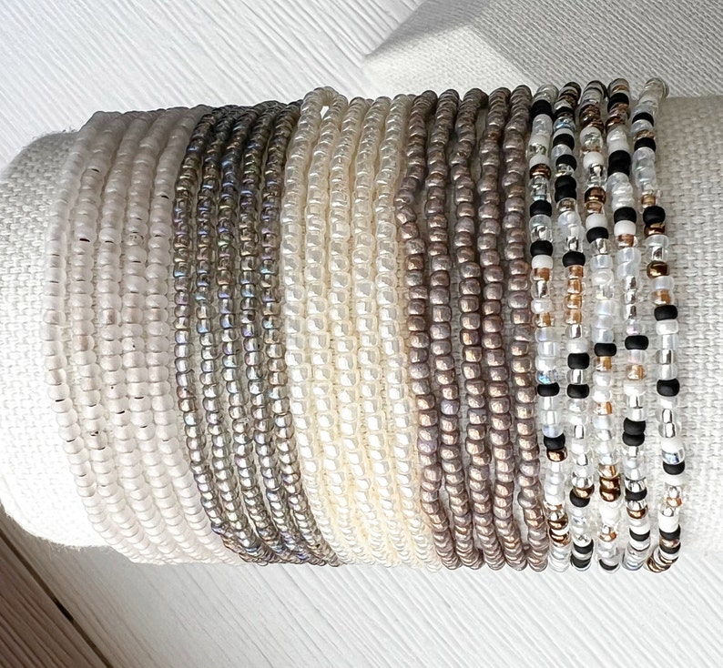 K&ouml;nnte beinhalten: Ein Stapel Perlenarmb&auml;nder in verschiedenen Wei&szlig;-, Grau- und Braunt&ouml;nen. Die Armb&auml;nder bestehen aus kleinen, runden Perlen und sind in einem abgestuften Muster angeordnet.