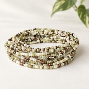 May include: A multi-strand bracelet composed of small, round glass beads in alternating shades of sage green, metallic bronze, and opaque white. The beads are strung on a flexible cord, creating a layered, circular stackable design.
