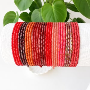May include: A collection of multiple thin beaded bracelets arranged in a gradient color pattern, transitioning from deep burgundy and dark red to bright orange, vibrant red, coral, and light pink, featuring small round glass beads on elastic cord.
