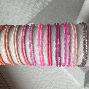 May include: A collection of thin, circular bracelets made from small, uniform glass seed beads in a gradient of colors ranging from coral and peach to various shades of pink, magenta, and a single metallic bronze strand arranged in a row.
