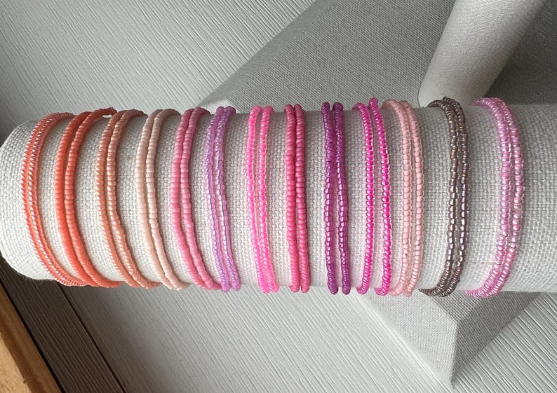 May include: A collection of thin, circular bracelets made from small, uniform glass seed beads in a gradient of colors ranging from coral and peach to various shades of pink, magenta, and a single metallic bronze strand arranged in a row.