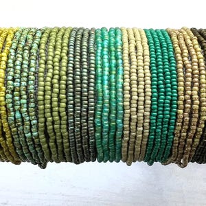 May include: A collection of multiple thin bracelets made from small, round glass seed beads in a gradient of colors including yellow, lime green, olive, dark green, turquoise, cream, emerald, tan, and dark brown, arranged in a side-by-side row.