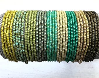 Puede incluir: Una colección de múltiples pulseras finas hechas de pequeñas cuentas de vidrio redondas en un degradado de colores que incluye amarillo, verde lima, oliva, verde oscuro, turquesa, crema, esmeralda, beige y marrón oscuro, dispuestas en fila.