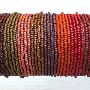 May include: A collection of multiple beaded bracelets arranged in a gradient color pattern. The beads transition through shades of dark brown, bronze, metallic pink, copper, deep purple, bright orange, and various tones of red in a linear sequence.