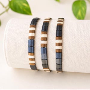 May include: Three bracelets composed of rectangular beads in alternating colors of metallic dark gray, bronze, and cream. The beads are arranged in a repeating pattern along each strand, creating a uniform and structured appearance for each piece.