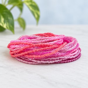 May include: A coiled stack of multiple thin bracelets made from small, round glass beads in various shades of pink, ranging from light pastel and bubblegum pink to vibrant magenta and deep berry tones, arranged in a circular, layered formation.