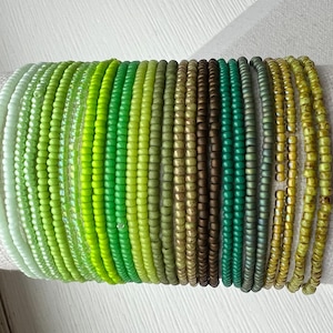 May include: A collection of twenty-one individual beaded bracelets arranged in a row. The bracelets feature a gradient of green shades, ranging from pale mint and lime to vibrant emerald, olive, forest green, and earthy brown tones.