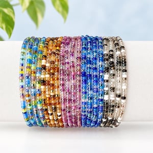 May include: A collection of multiple thin bracelets made from small, round, multicolored glass beads. The bracelets feature a gradient of colors including shades of blue, green, yellow, orange, brown, pink, purple, and black, white, and clear.
