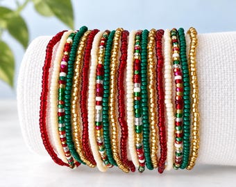 Puede incluir: Una colección de quince pulseras de cuentas individuales dispuestas en fila. Las pulseras presentan pequeñas cuentas de vidrio redondas en una paleta de colores festivos de oro metálico, verde bosque profundo, rojo cereza brillante y blanco crema opaco.