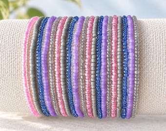 Puede incluir: Un conjunto de múltiples pulseras elásticas hechas de pequeñas cuentas de vidrio redondas en una paleta de colores repetitiva de rosa, morado claro, azul real y plata metálica, dispuestas en una fila vertical sobre una tela texturizada de color crema.