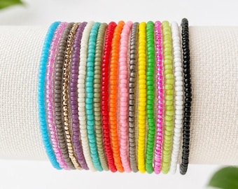 May include: A collection of sixteen thin beaded bracelets arranged in a row, featuring a vibrant spectrum of colors including blue, pink, grey, metallic gold, purple, white, teal, green, red, orange, yellow, lime, and black glass seed beads.