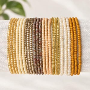 May include: A collection of eighteen individual beaded bracelets arranged in a row. The bracelets feature a variety of small, round beads in shades of gold, cream, beige, speckled brown, peach, white, and mustard yellow, creating a gradient effect.