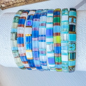 May include: A collection of colorful beaded bracelets. The bracelets feature a variety of rectangular beads in shades of blue, green, white, and gold. The bracelets are stacked together, showcasing their design and color variations. The beads appear to be approximately 0.25 inches in length.