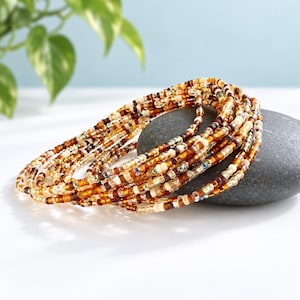 May include: A multi-strand bracelet composed of small glass seed beads in various shades of amber, brown, cream, and clear. The beads are strung on a flexible cord, creating a layered, circular arrangement that rests upon a smooth gray stone.