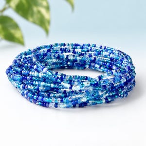 May include: A multi-strand bracelet composed of numerous small glass beads in various shades of blue, including navy, royal blue, turquoise, and clear crystal. The beads are arranged in a circular, coiled formation with a consistent texture.