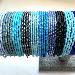 May include: A collection of colorful beaded bracelets in shades of blue, gray, and white. The bracelets are arranged in a row and are displayed on a gray fabric background.