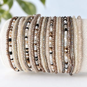 May include: A stack of multiple thin bracelets made from small seed beads in a neutral color palette. The beads feature shades of cream, beige, metallic bronze, translucent white, and matte black, arranged in various repeating patterns and solid rows.