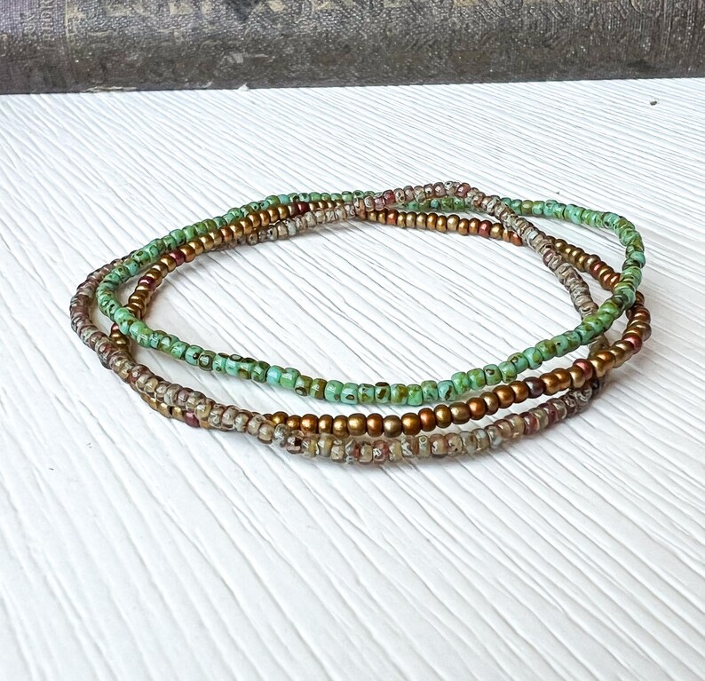 May include: Three layered beaded bracelets in shades of green, gold, and brown. The bracelets are made of small, round beads.